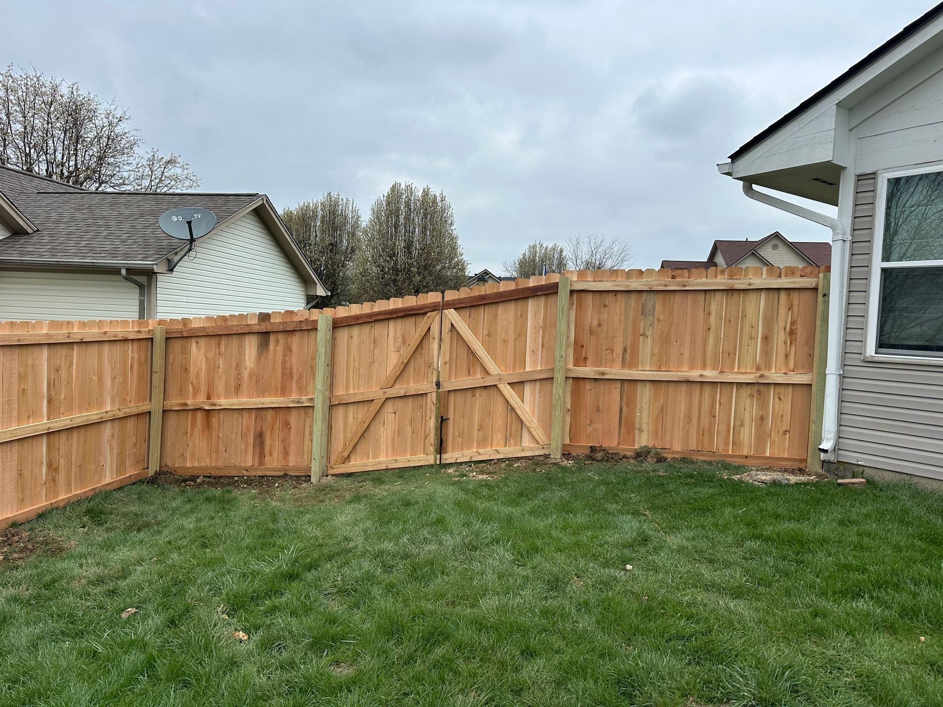 Wooden fence with gate in a grassy backyard, adjacent to a house, cloudy sky.