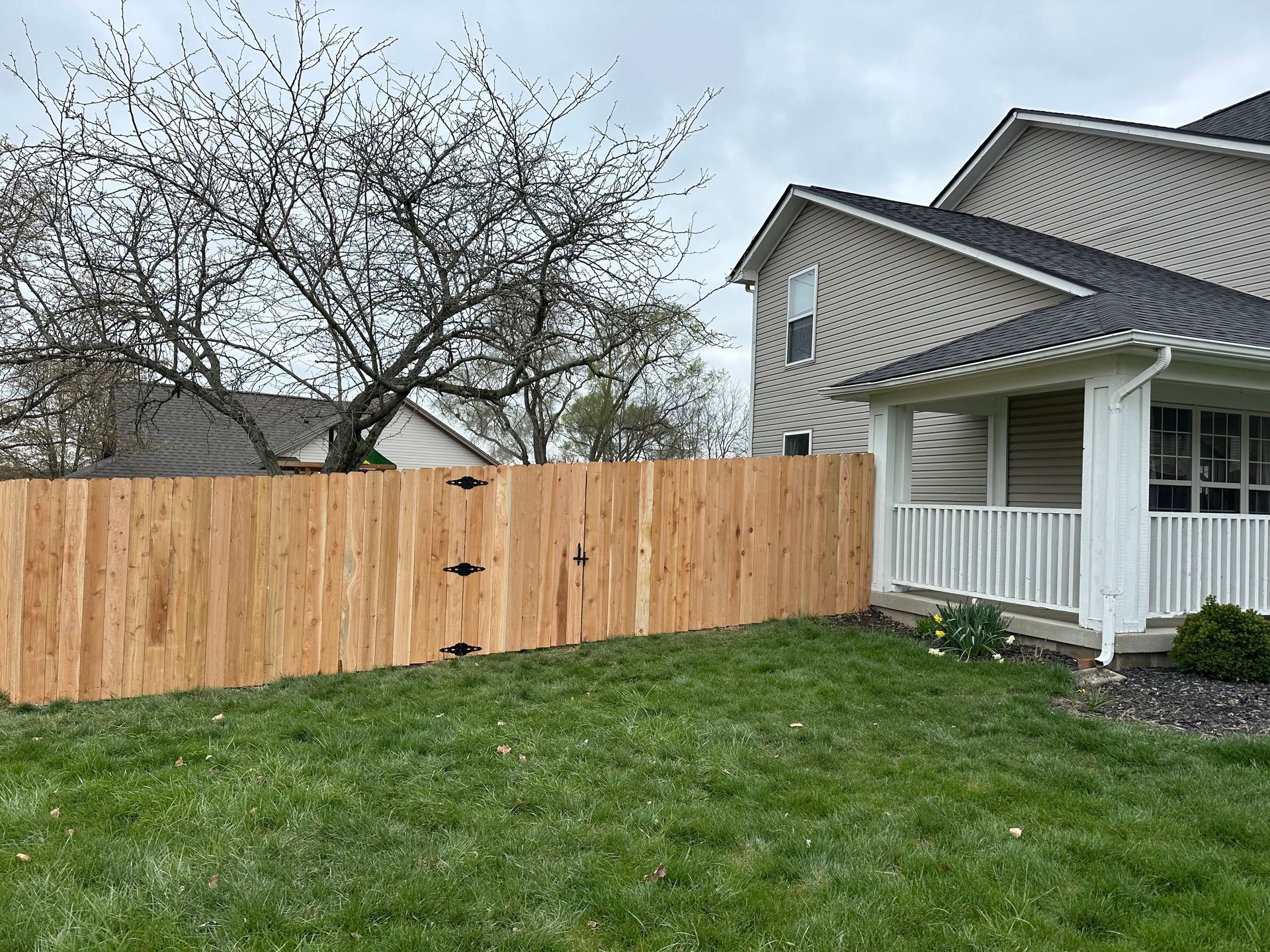 Wooden fence in a yard next to a two-story beige house and porch with white railings, under a cloudy sky.