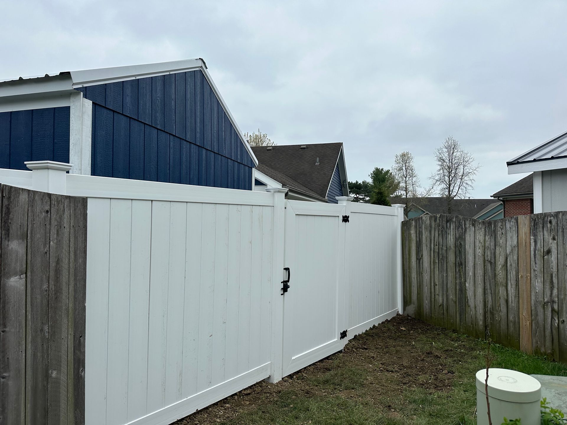 White vinyl fence with gate, bordering a wooden fence and a blue-sided building under a cloudy sky.