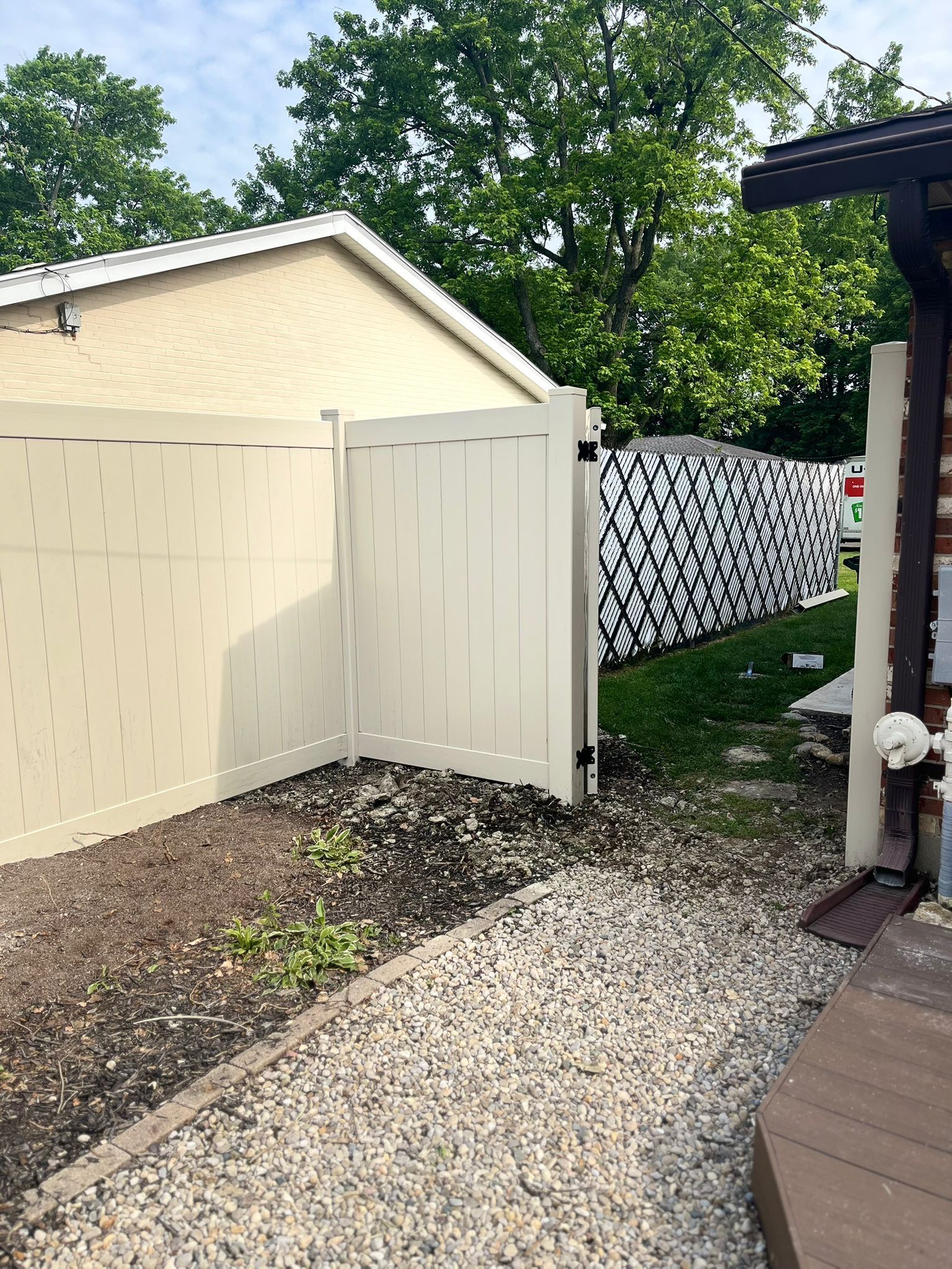 Beige vinyl fence with a gate, next to a building and gravel path. Black and white patterned fence in the distance.