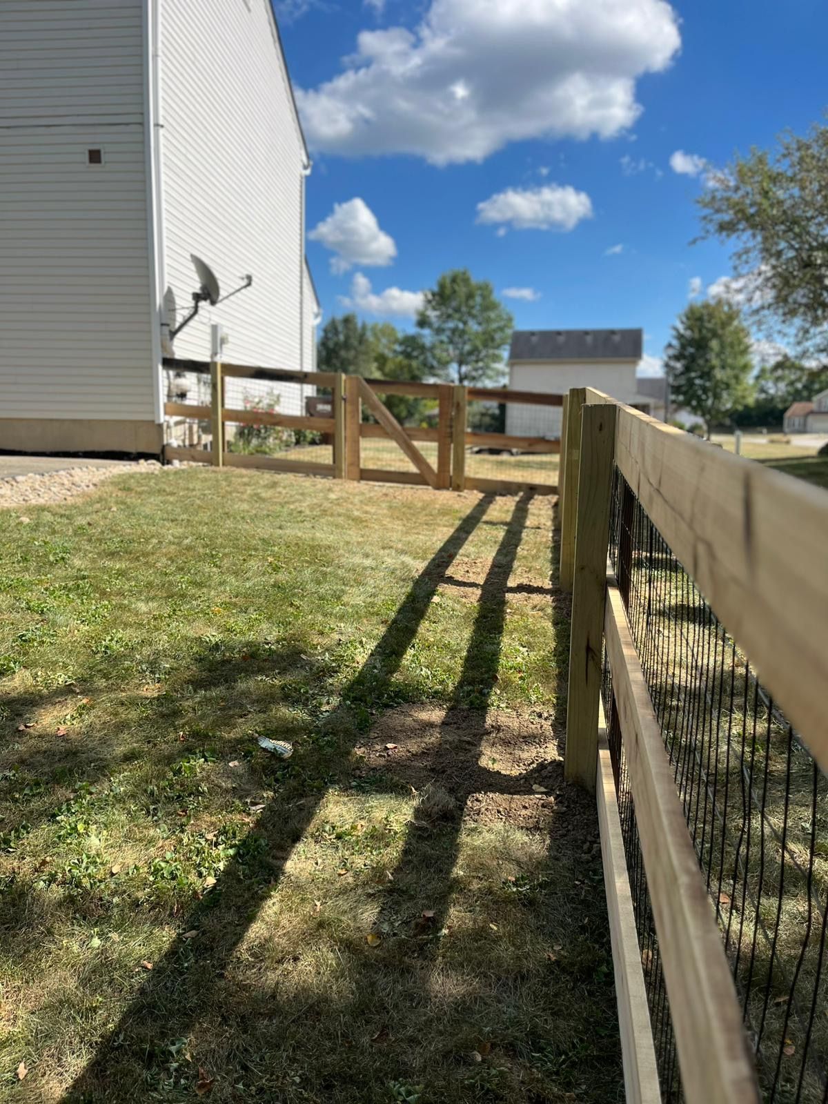 A newly built wooden fence with a gate encloses a grassy area next to a white building.