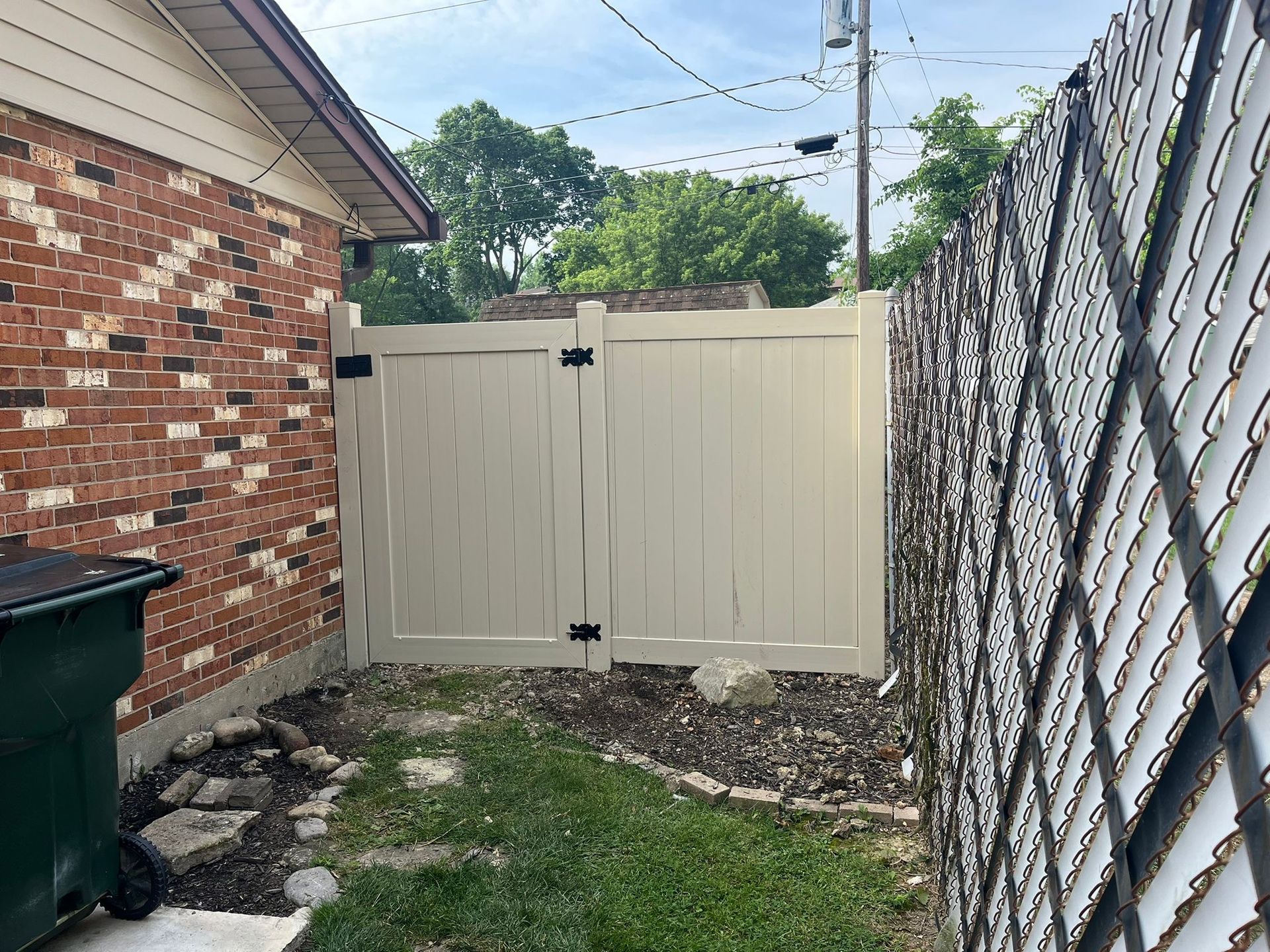 A beige gate in a backyard between a brick wall and a chain-link fence.