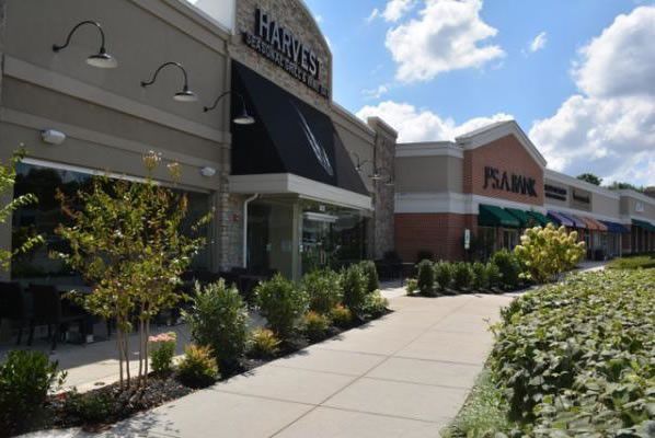 Shopping center storefronts with awnings, landscaping, and a wide sidewalk under a sunny sky