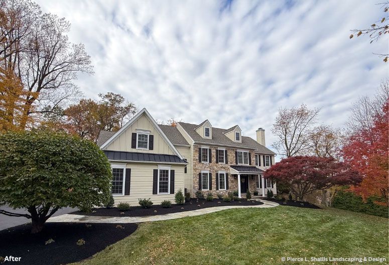 Large suburban house with beige siding, black shutters, and autumn trees under a cloudy sky