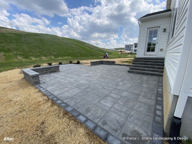 Stone patio beside a white house with steps, retaining wall, and grassy hillside under a cloudy sky