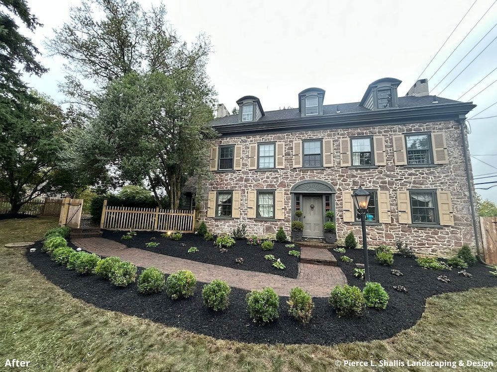 Stone house with dark shutters and dormer windows, fronted by a curved landscaped garden and trees