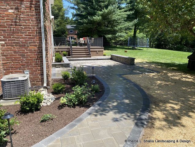 Curved stone walkway beside brick house, landscaped garden beds, and lawn with trees.