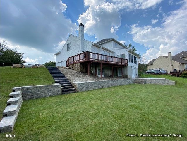White two-story house with a deck, stone retaining wall, and broad grassy backyard under a cloudy sky