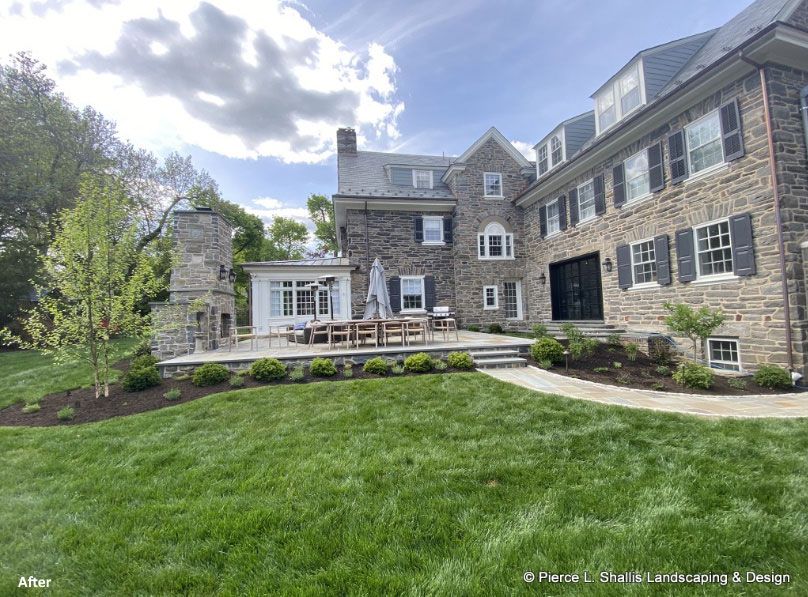 Stone house with a deck and lawn, featuring large windows and a landscaped backyard under a cloudy sky