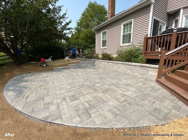 Paver patio beside a house and wooden deck, with workers in the background.