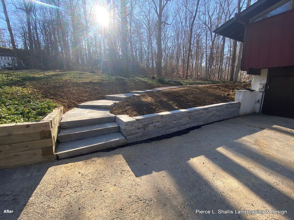Driveway with concrete steps, retaining wall, bare trees, and bright sunlight beside a red building