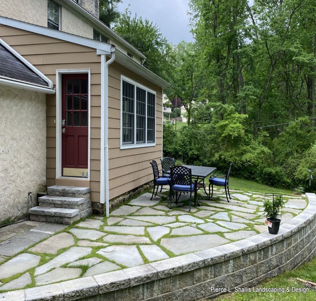 Small stone patio beside a tan house, with blue chairs, table, red door, and potted plants under trees.