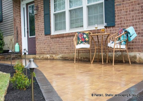 Rainy brick patio with wicker chairs, colorful cushions, and a small table beside a house window.