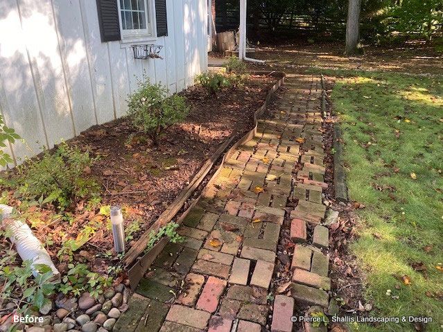 Brick garden path beside a white house, bordered by mulch and grass, with some uneven bricks and a small window