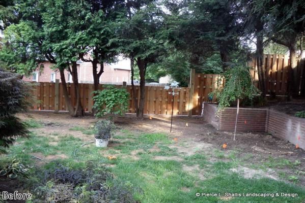 Backyard with grass, trees, wooden fence, and a brick retaining wall at dusk