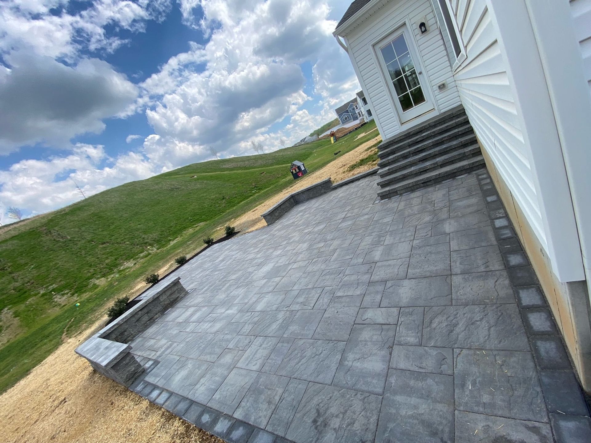 Stone patio beside a white house, with steps and grassy hillside under a cloudy sky