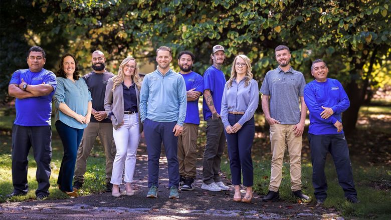 Group of nine people standing outdoors in front of leafy trees, posing for a photo.