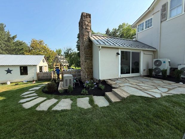 Backyard patio with stone path, small white shed, and stone chimney beside a house