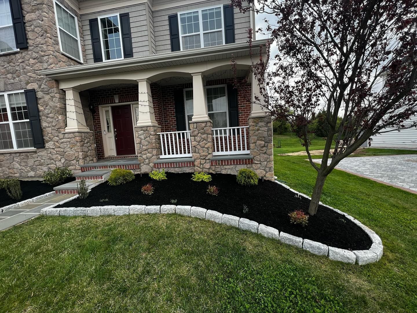 Brick house front porch with white columns, curved stone edging, mulch bed, and a small tree.