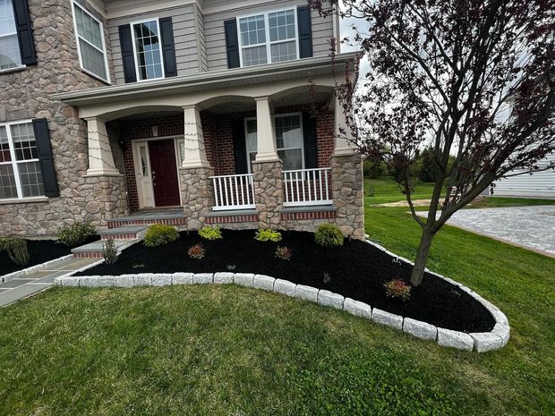 Brick house front porch with white columns, curved stone edging, mulch bed, and a small tree.