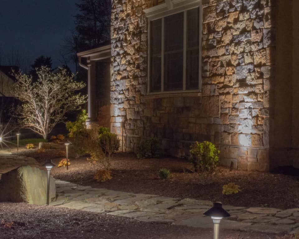 Lit stone house exterior at night with a garden path, shrubs, and warm landscape lighting