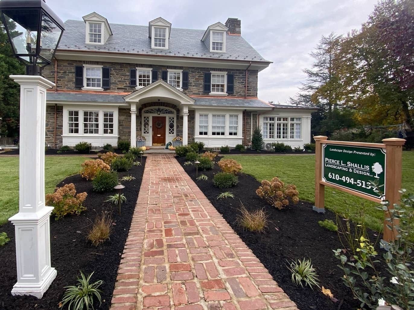 Historic stone house with brick walkway, manicured landscaping, and a green sign in front