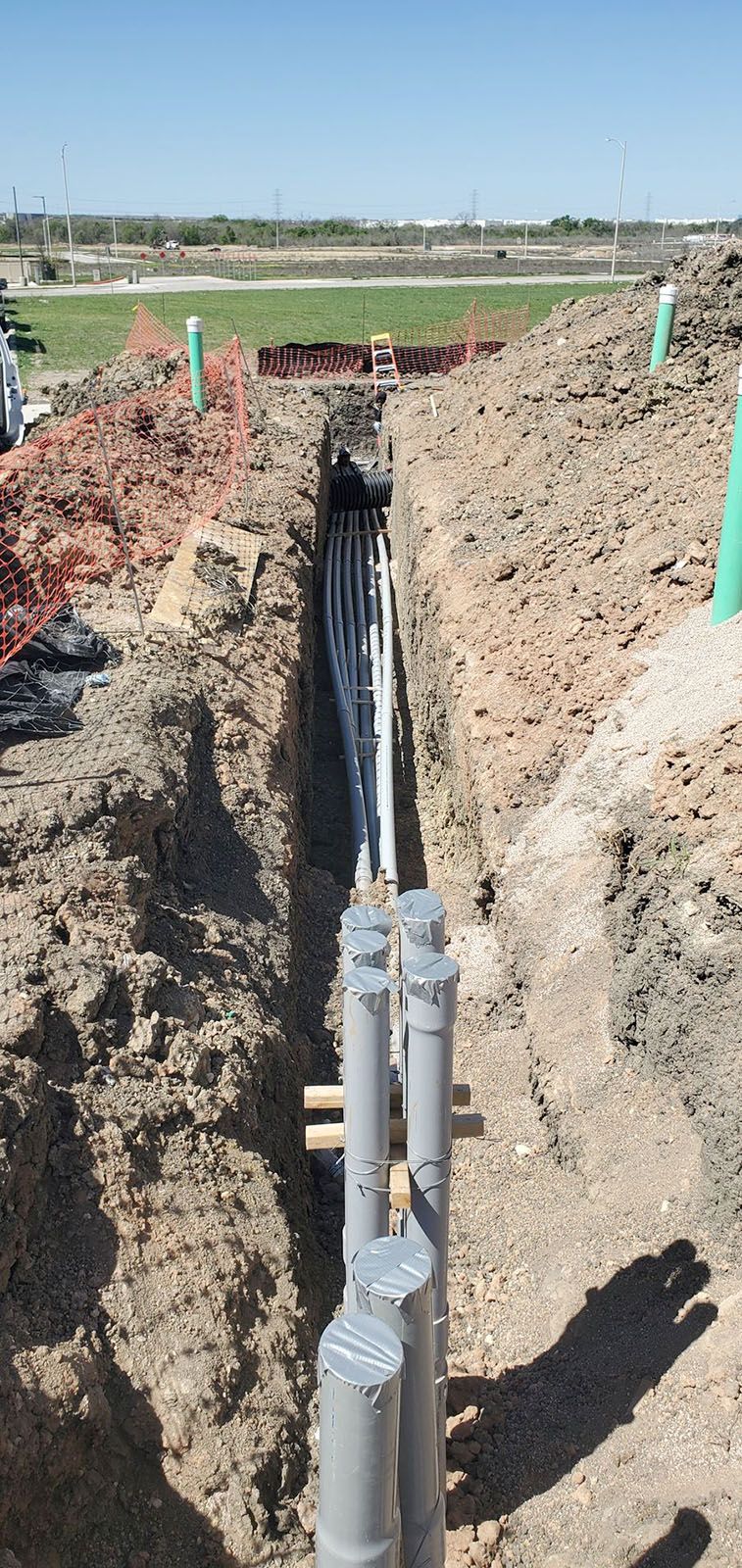 A deep trench in a construction site containing multiple underground utility conduits and vertical PVC pipes