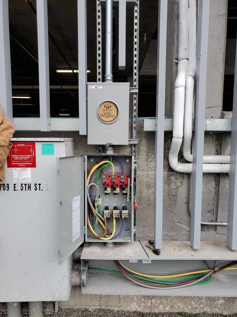 A electrical box is sitting on the side of a building next to a fence.