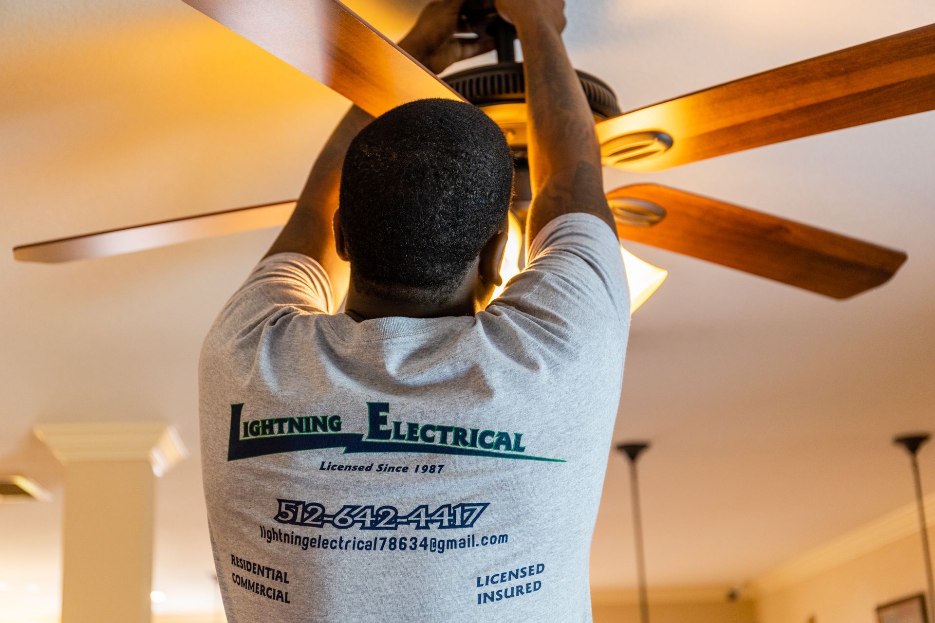 A man in a lightning electrical shirt is fixing a ceiling fan.