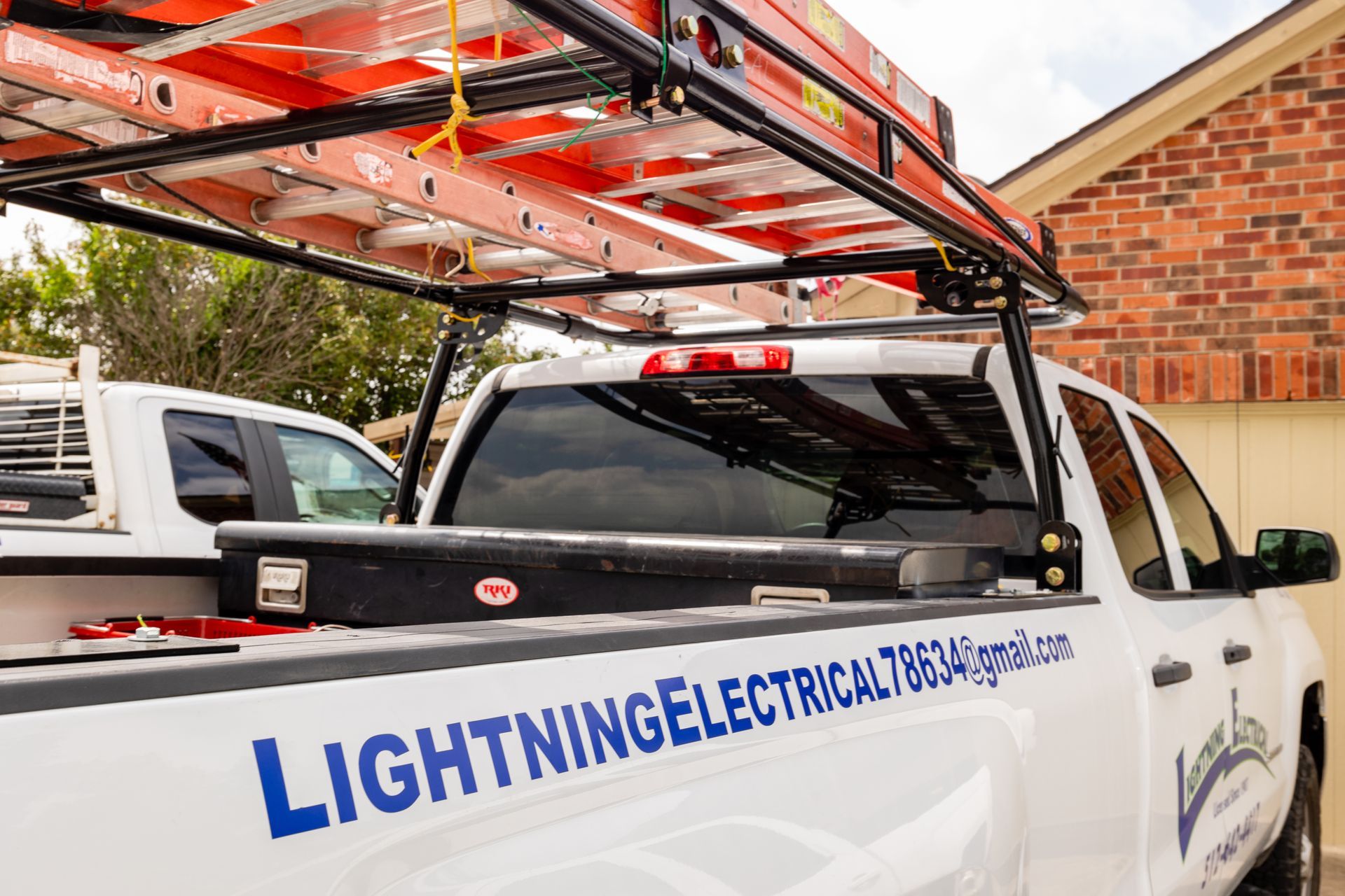 A white truck with a ladder rack on the back is parked in front of a brick building.