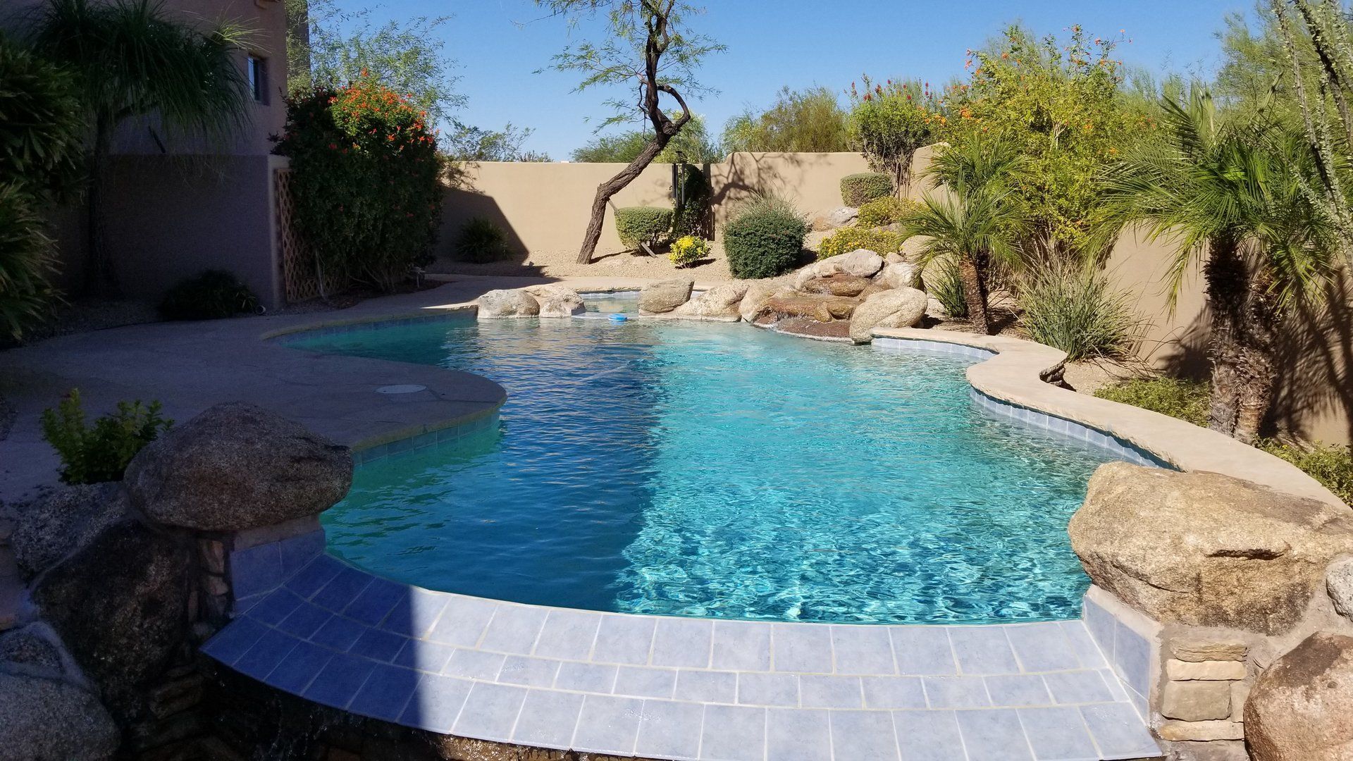 Pool with blue water surrounded by stone and landscaping, featuring a small waterfall.