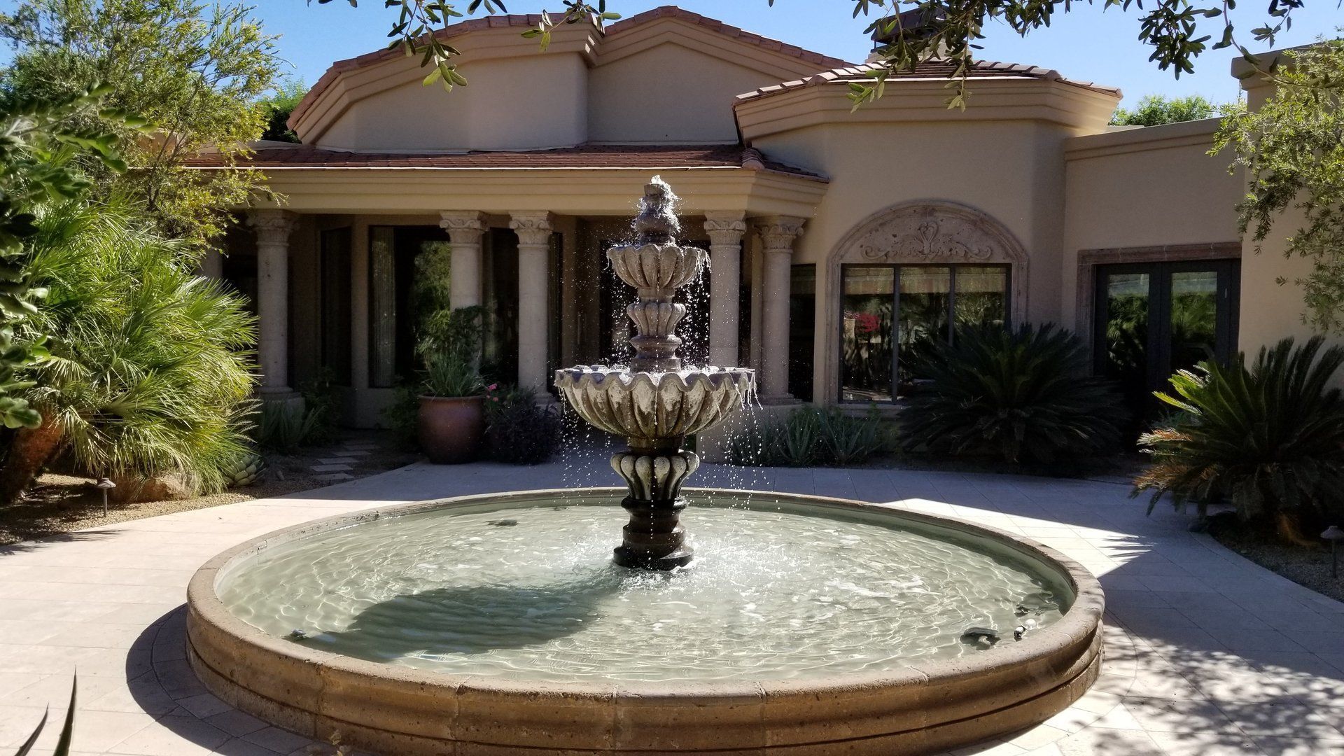 Fountain in a round basin in front of a beige stucco house with columns and windows, palm trees around.