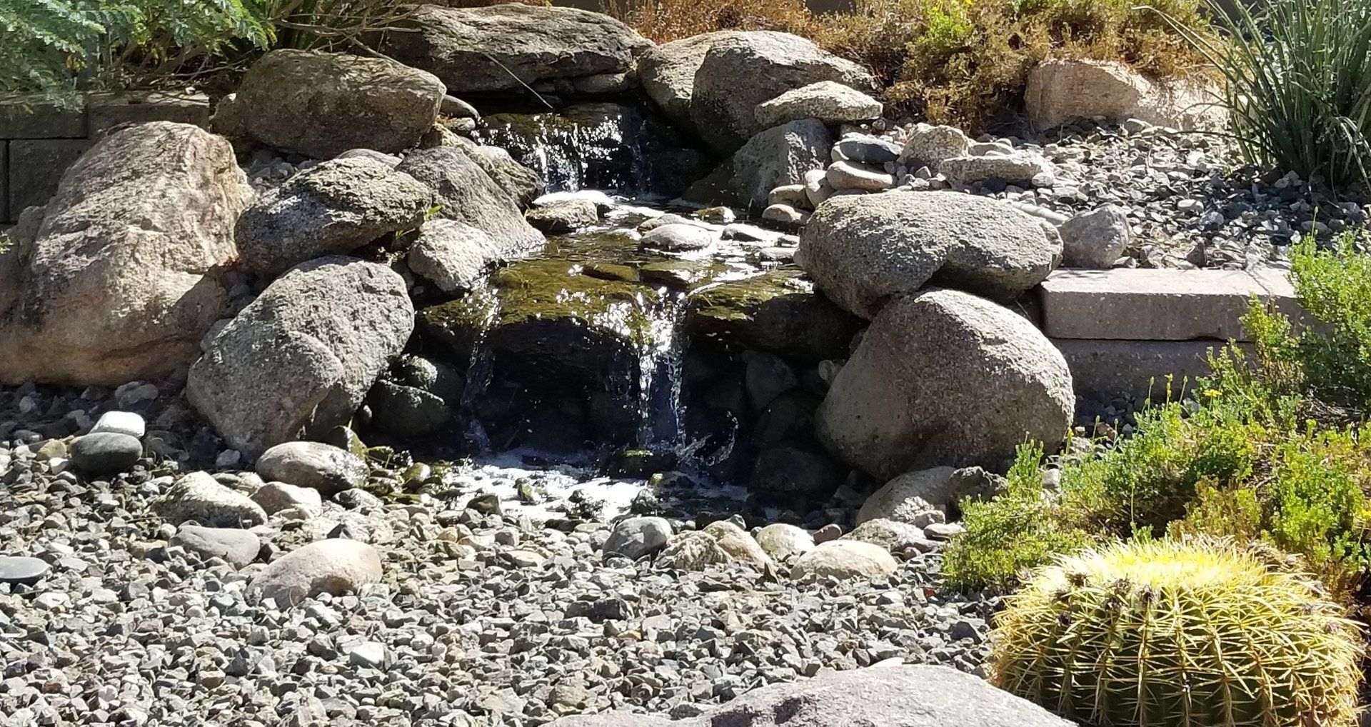 Small waterfall cascading over rocks in a garden, with gravel, boulders, and plants.