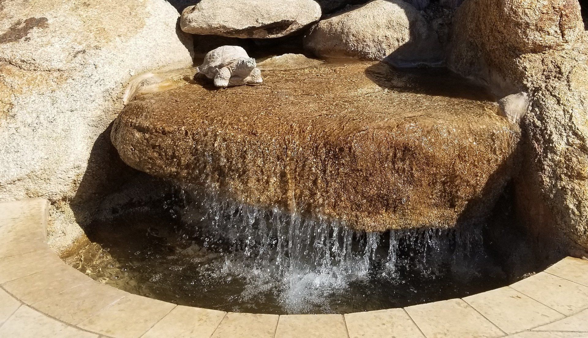 Water cascades from a rock fountain into a small basin; beige stone.