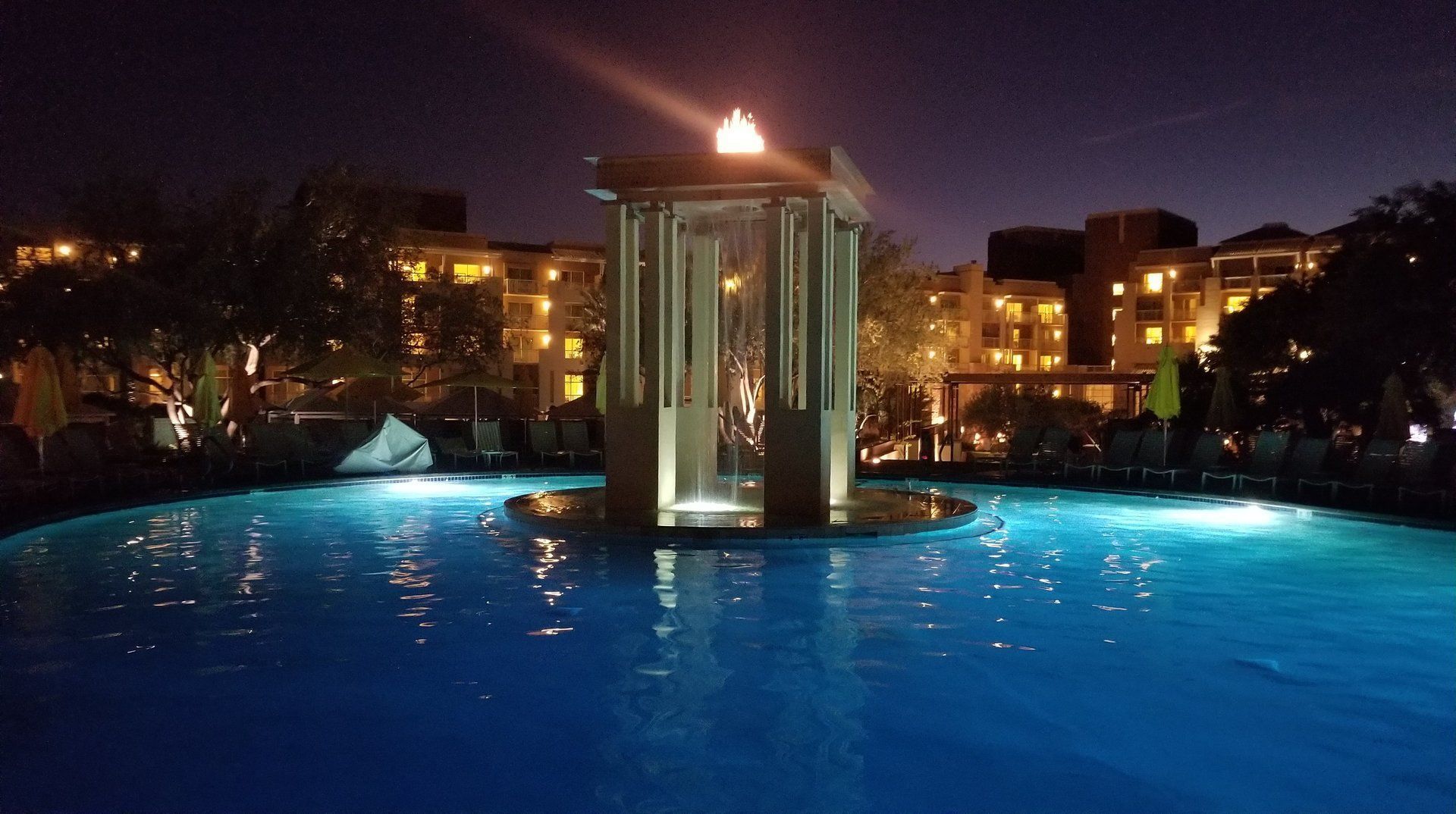 Nighttime scene: illuminated fountain in a pool, buildings in background, dark blue sky.