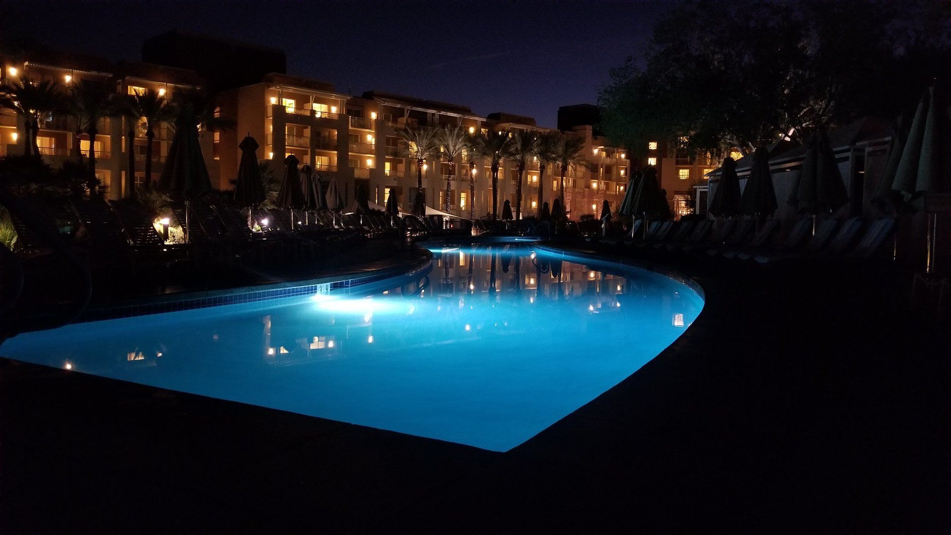 Nighttime view of a resort pool, reflecting lights from the building and palm trees.