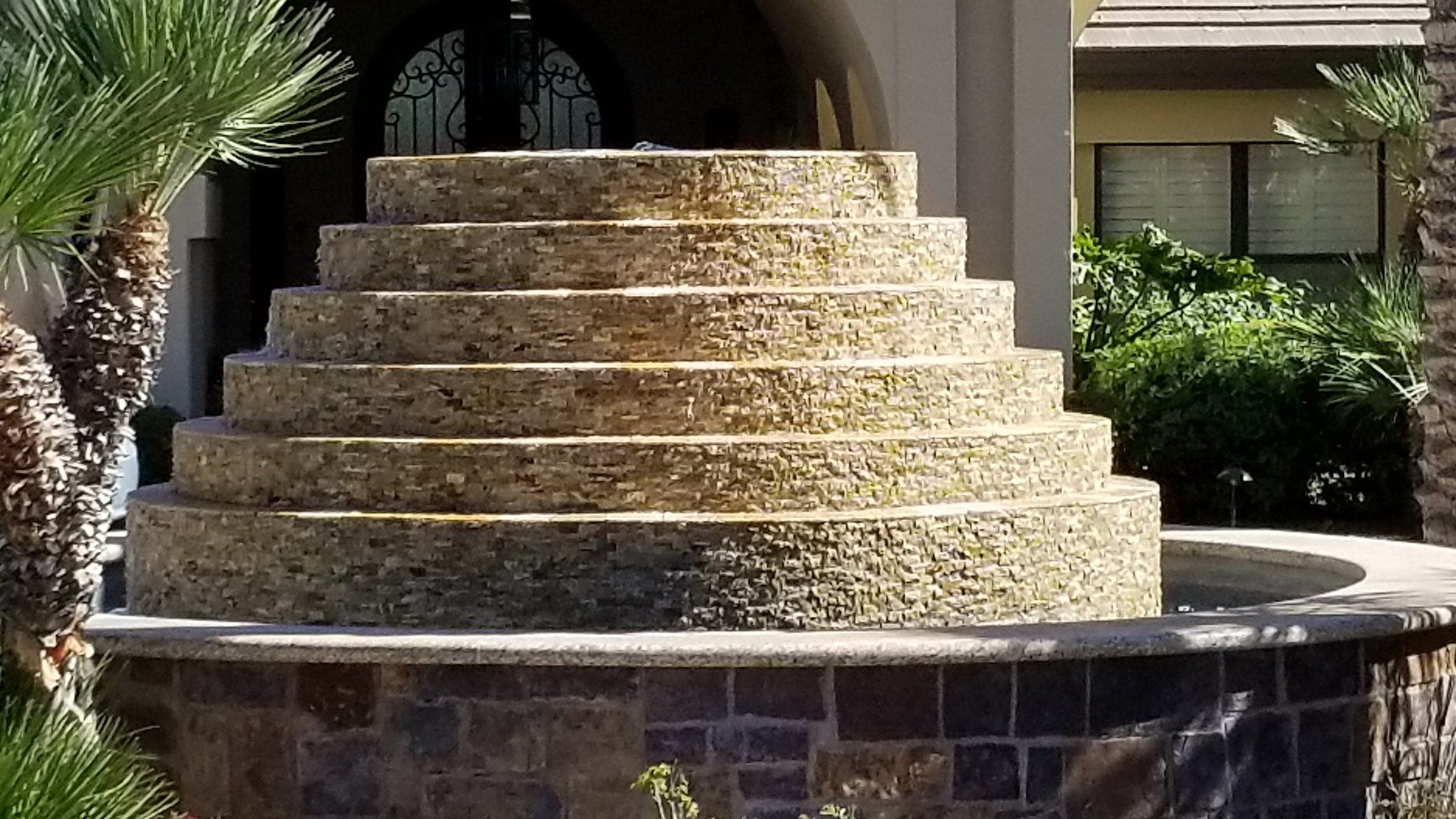 Tiered stone water fountain in front of a building, with greenery nearby.