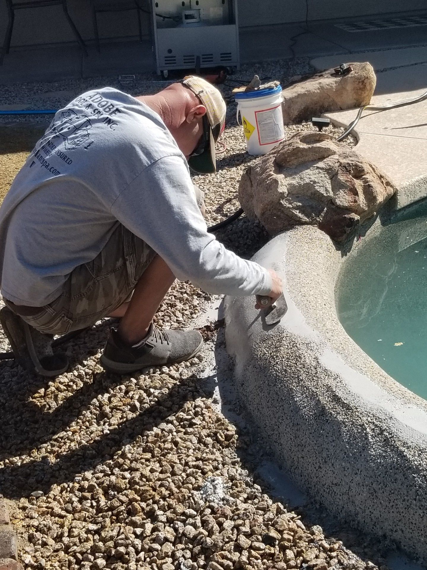 A man working on the edge of a pool, applying sealant. Gravel and rocks surround the pool.