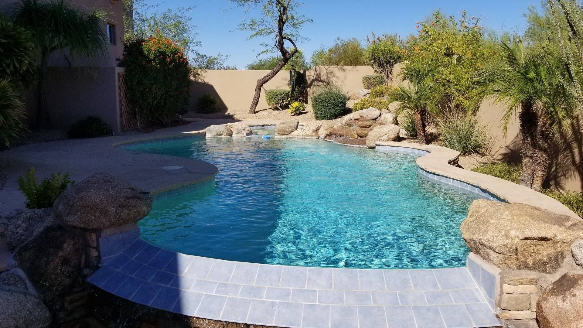 Swimming pool with blue water surrounded by rocks, trees, and a stone bridge.