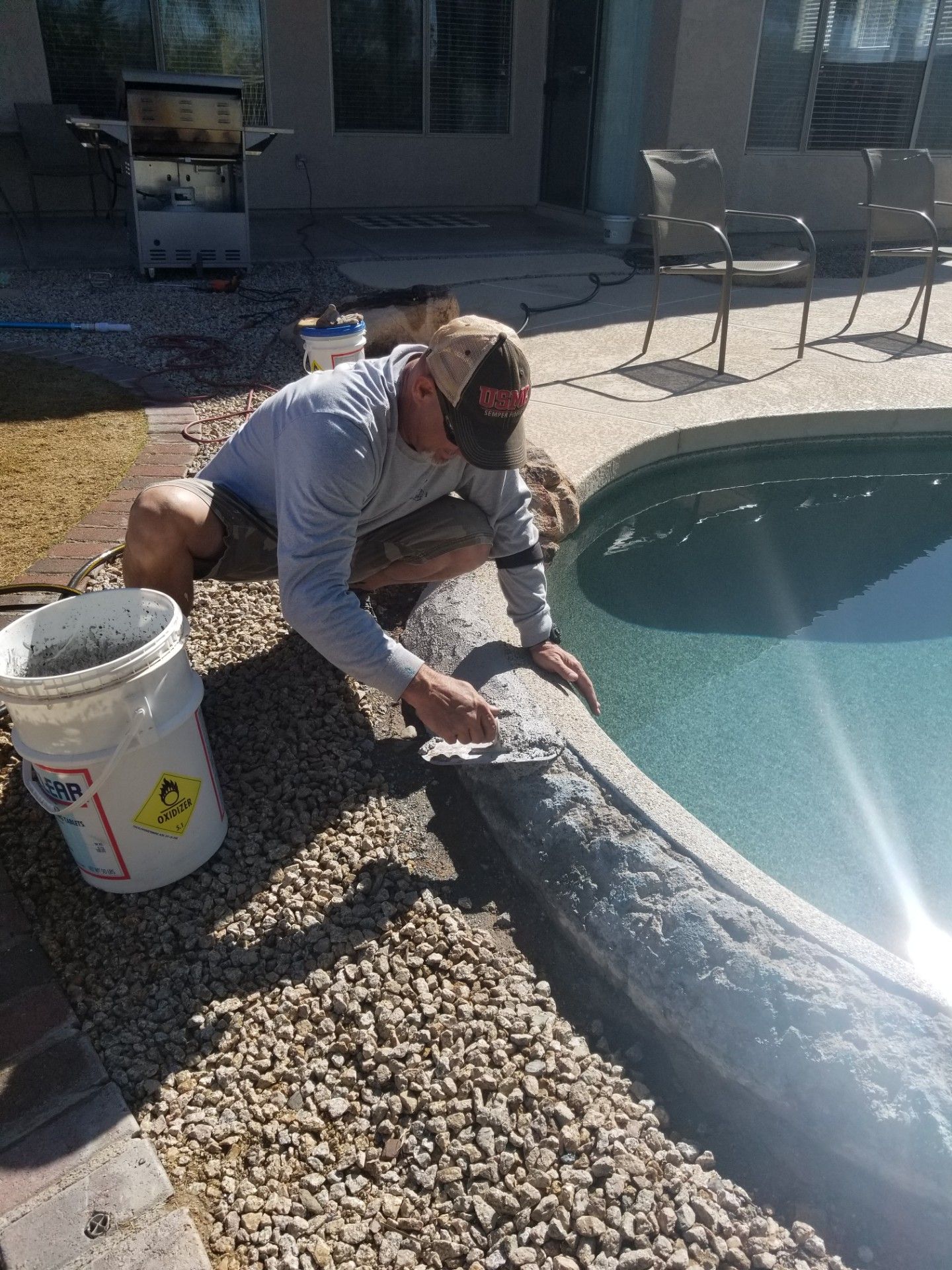 Man repairs pool coping with a trowel, near water. He's crouching with bucket and small gravel surrounding.
