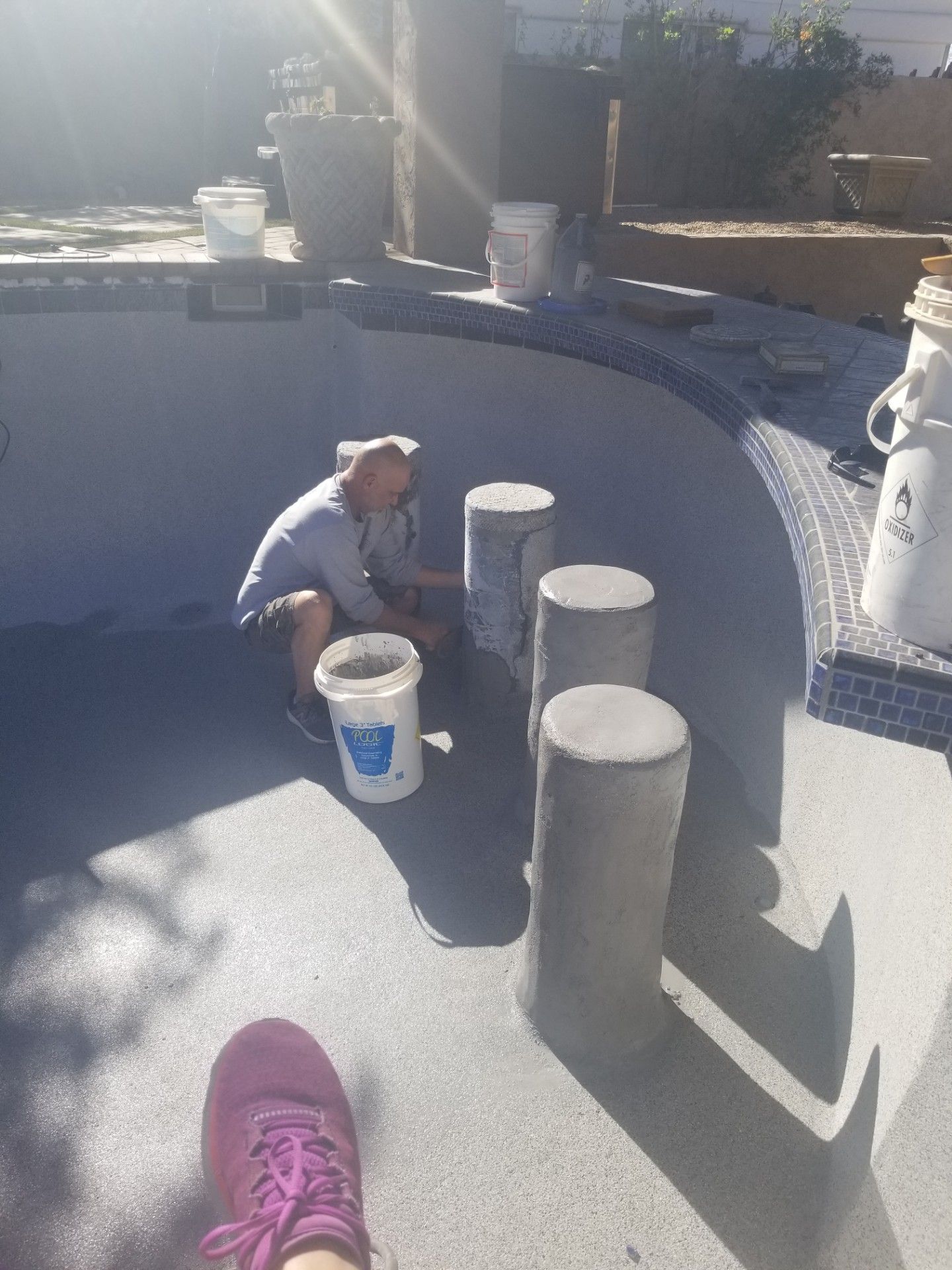 Man working on a pool, applying material to concrete pillars. Buckets nearby. Sunny outdoor setting.