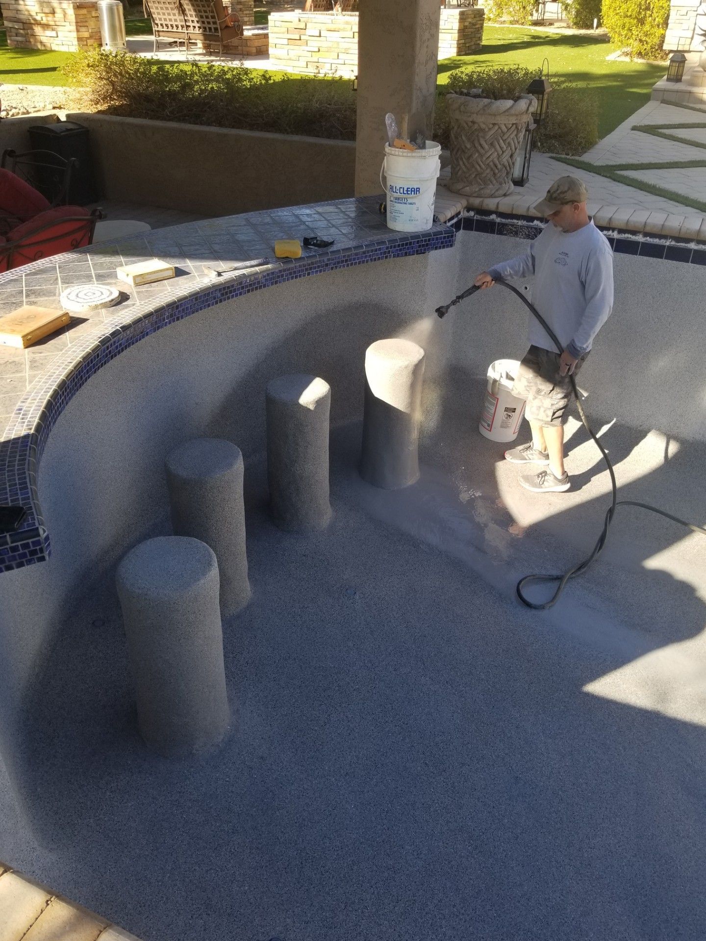 Man sprays a pool interior with resurfacing material. Gray pool with bar and concrete stools. Sunny outdoor setting.
