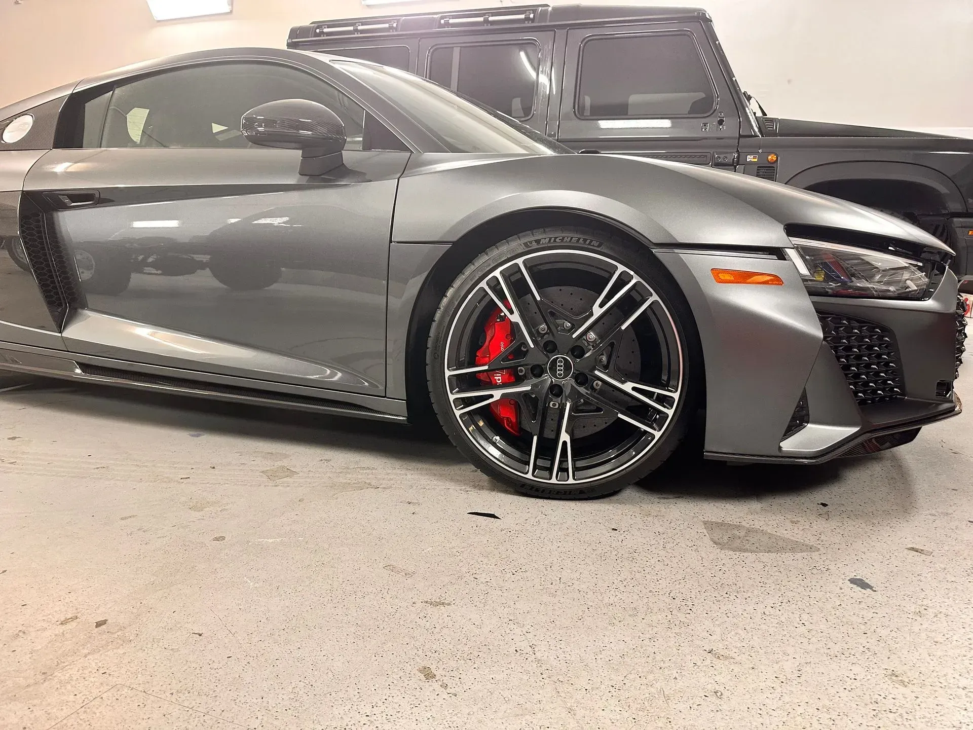 Gray Audi R8 sports car with red brake calipers, parked next to a black SUV in a garage.