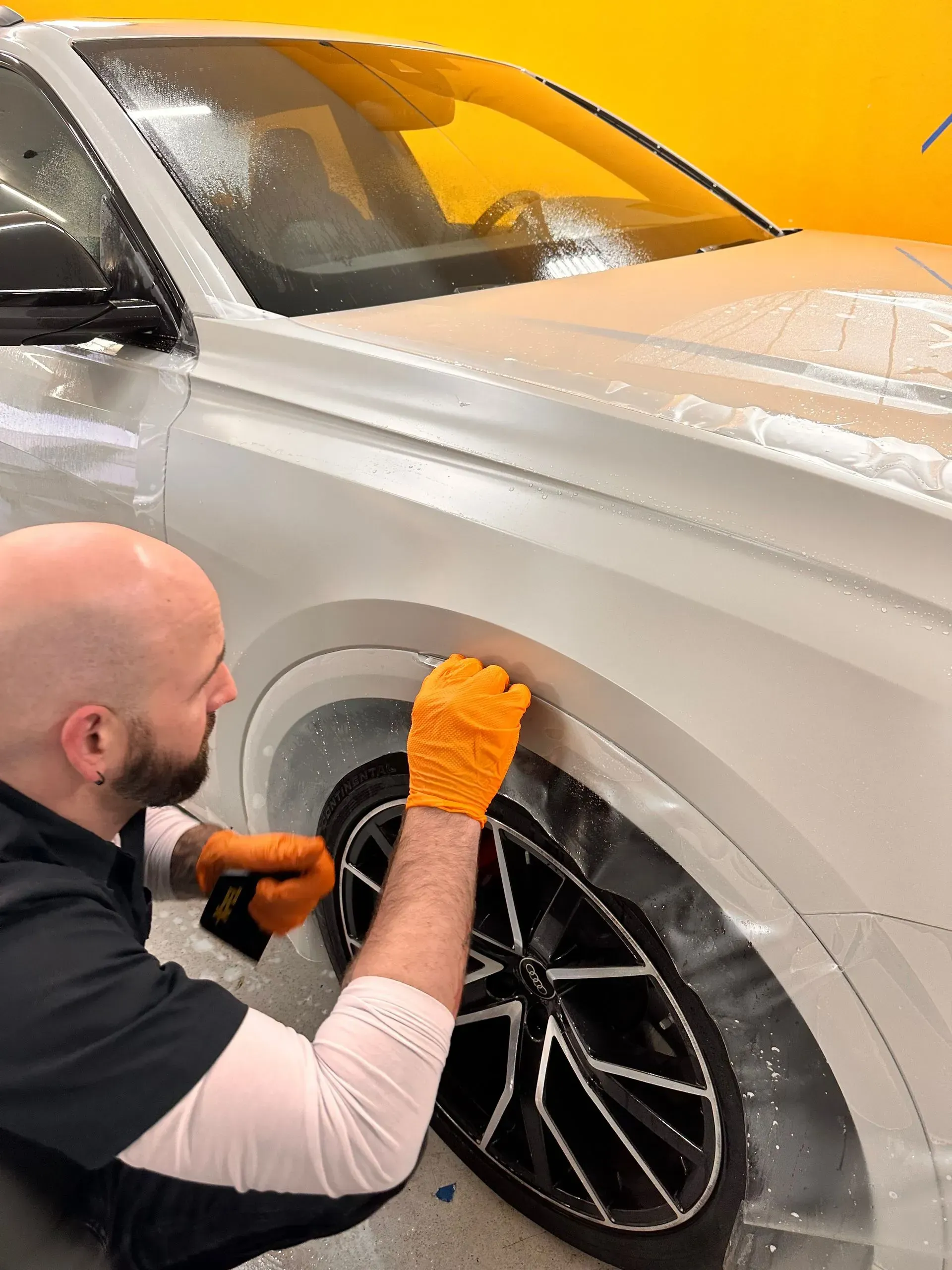 A person applying protective film to the front fender of a white car, wearing gloves.