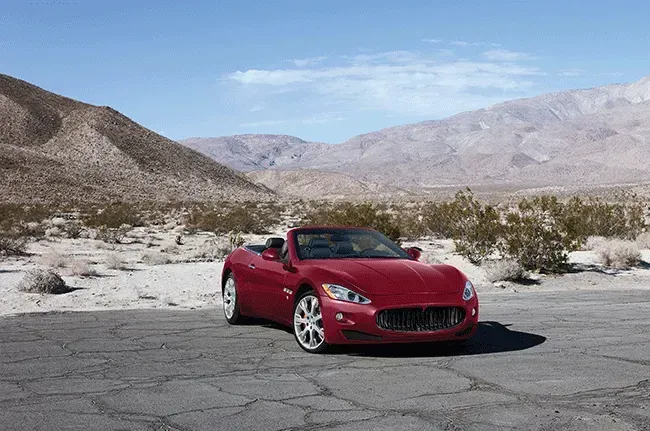 Red convertible car parked on a cracked road, desert landscape with mountains in the background.