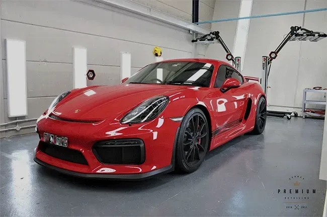 Red Porsche Cayman in a garage, black wheels, lights shining, gray floor.
