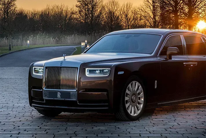 Brown Rolls-Royce Phantom luxury sedan parked on a brick road with the sun setting in the background.