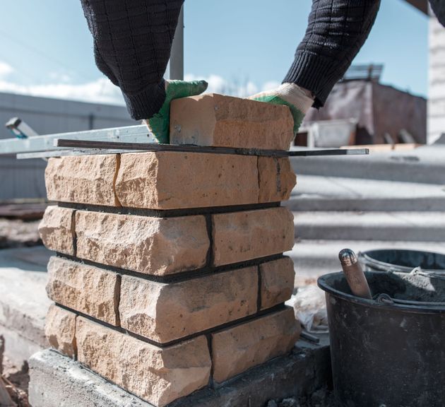 Hands wearing gloves placing a brick on top of a brick column under construction outdoors.