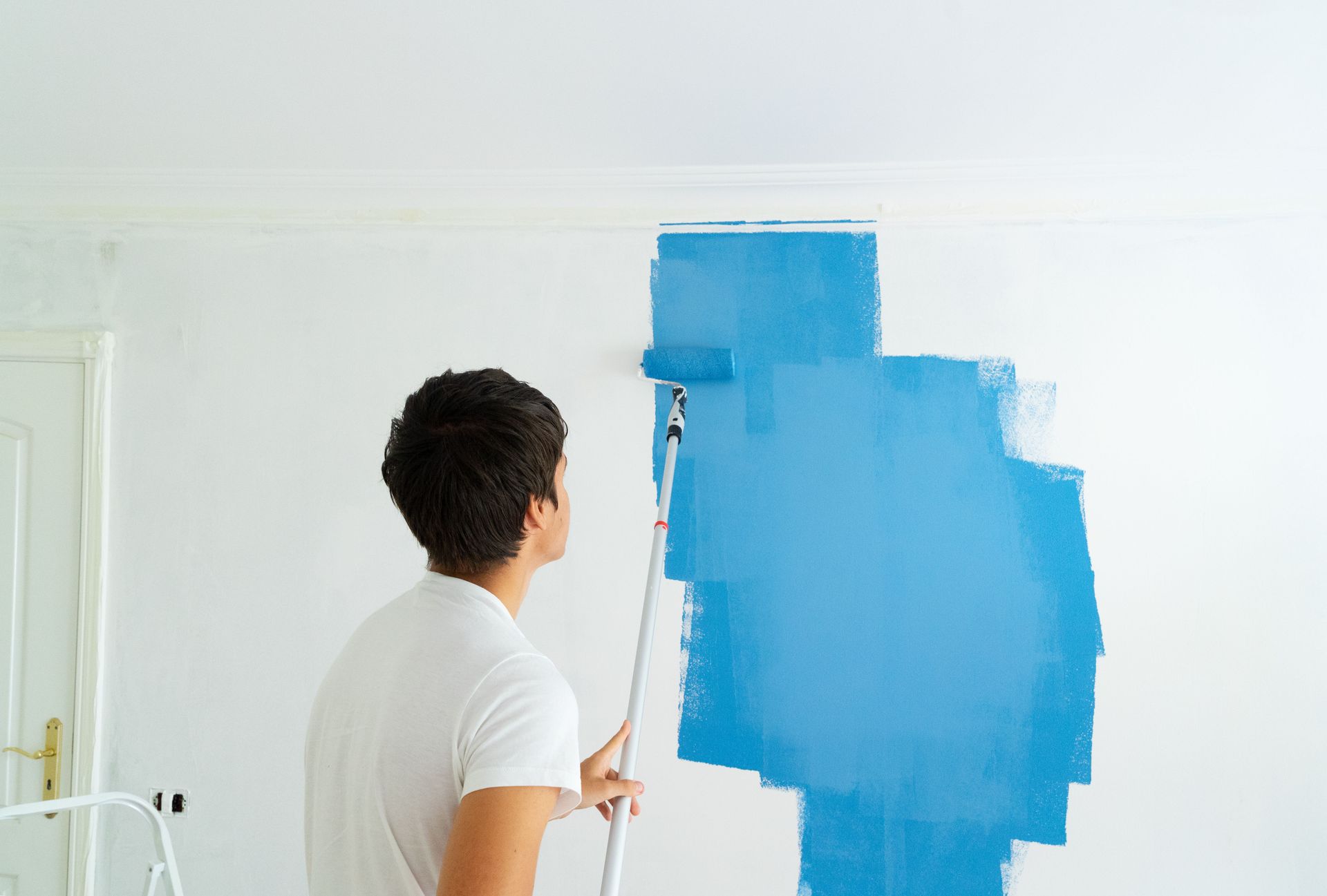 A person uses a paint roller on an extension pole to paint a white wall bright blue.