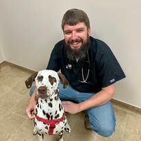 Smiling man kneels beside a Dalmatian in a clinic, both facing the camera.